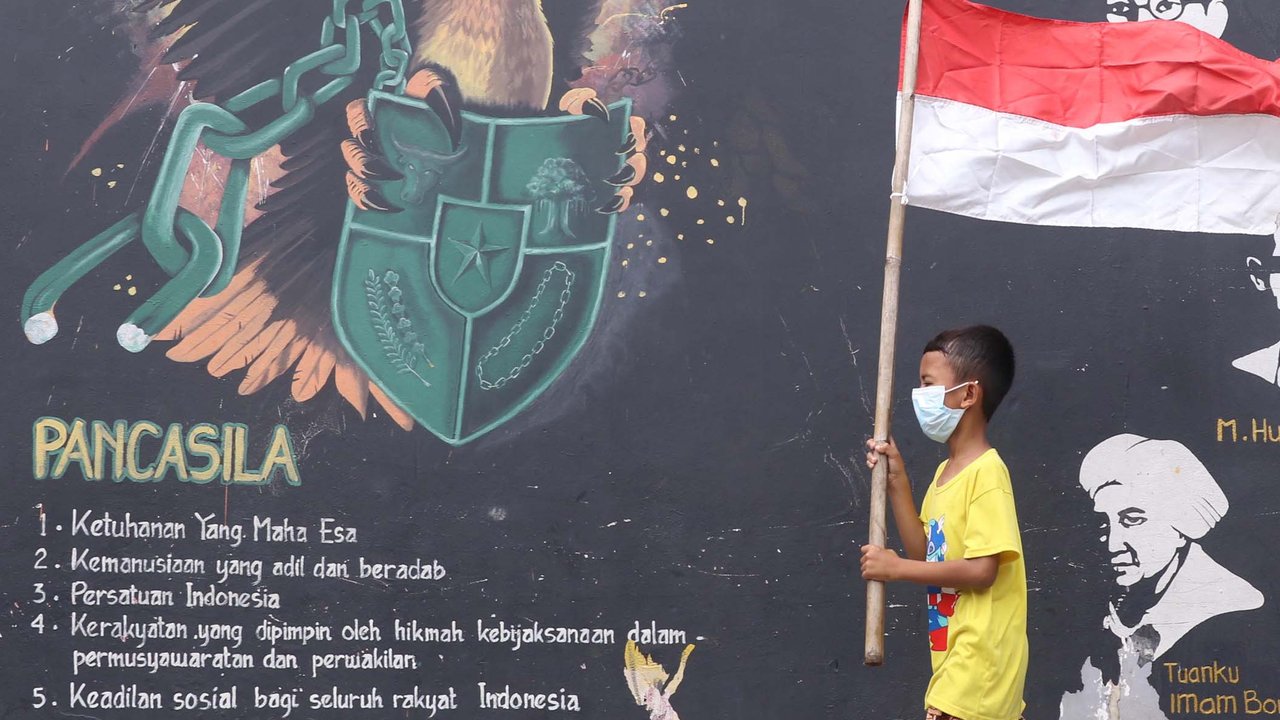 Anak-anak melintas membawa bendera merah putih berlatar belakang mural Pancasila di Kampung Pancasila, Karang Tengah, Kota Tangerang, Selasa (1/6/2021). Sejumlah kegiatan diadakan warga, Komunitas Taman Potret dan TNI ini untuk memperingati Hari kelahiran Pancasila. (Liputan6.com/Angga Yuniar)