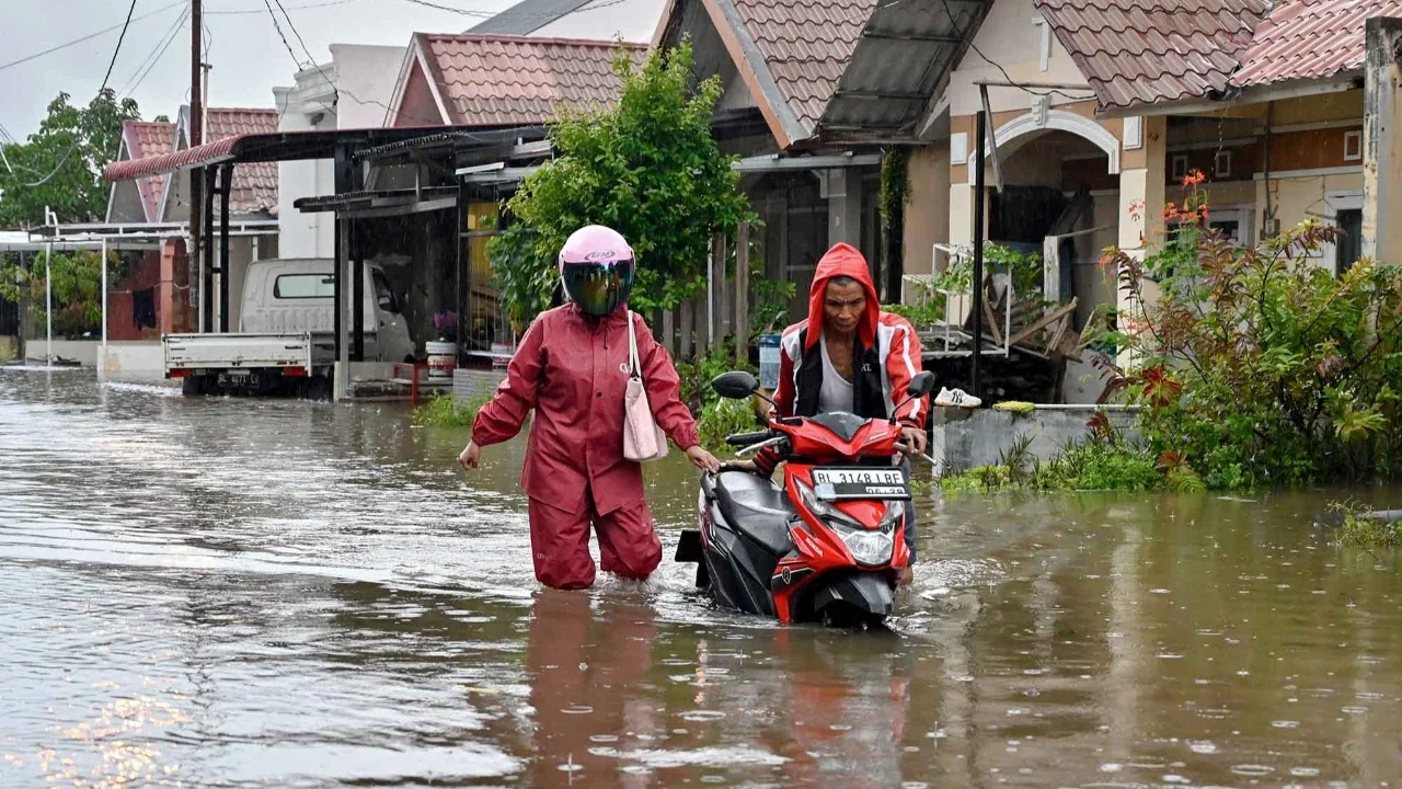 Warga mendorong sepeda motornya menerobos banjir di jalan yang tergenang setelah hujan deras di permukiman Darul Imarah, pinggiran Banda Aceh, Kamis (27/11/2025).