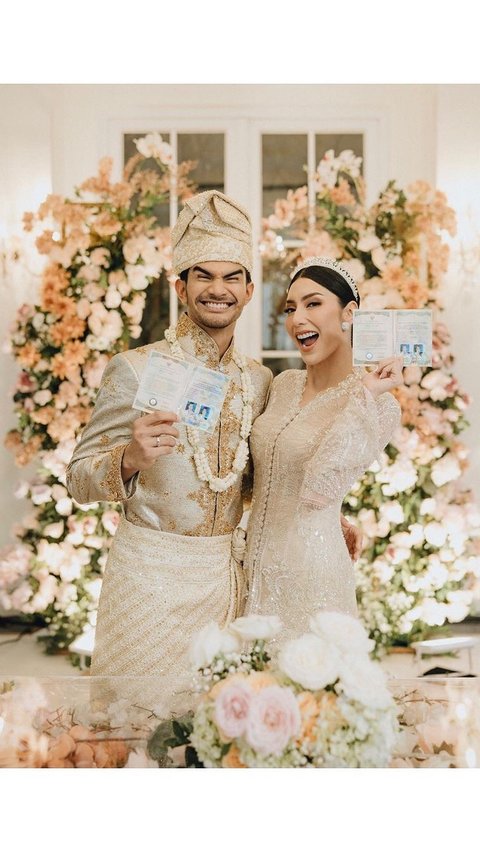 Pasangan ini tampak serasi mengenakan baju pengantin warna krem. Kedunya juga terlihat memamerkan buku nikah.
