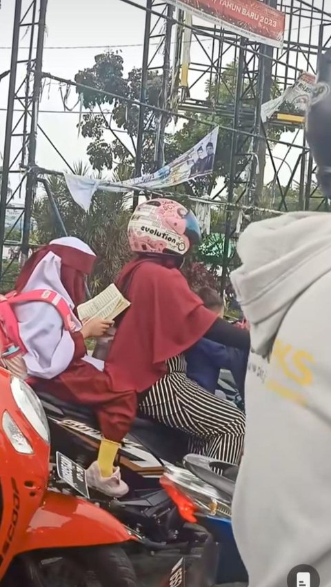 Sambil duduk di motor yang dikendarai ibunya, siswa SD tersebut tampak serius membaca Al-Qur'an.