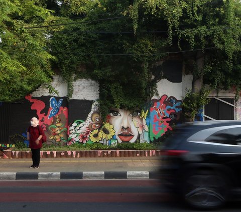 Seorang warga tampak sedang menunggu kendaraan umum di trotoar dengan latar belakang pemandangan mural karya petugas PPSU di kawasan Jalan Malaka, Jakarta.