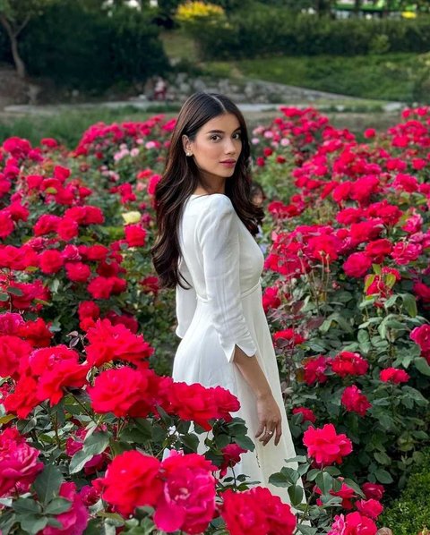 Sabrina mempesona dengan penampilan anggun dalam gaun putih, dikelilingi oleh kecantikan bunga mawar yang mekar di sekelilingnya.
