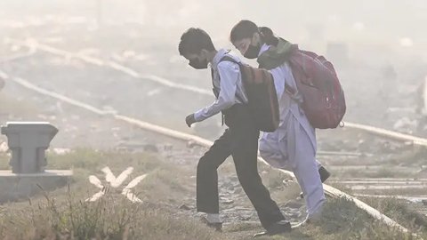 Anak-anak sekolah mengenakan masker berjalan melintasi rel kereta api di tengah kabut asap tebal di Lahore, Pakistan pada Rabu 20 November 2024. (Arif ALI/AFP)
