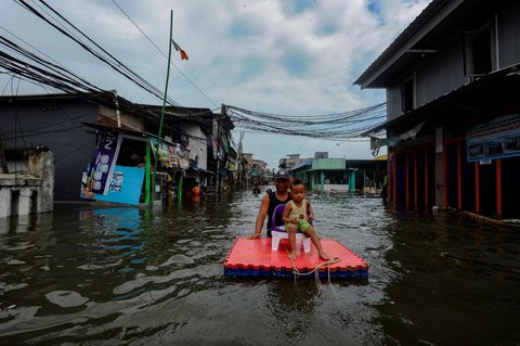 Seorang warga bersama anaknya menggunakan rakit sederhana saat melewati banjir rob yang melanda kawasan Muara Angke, Jakarta, Senin (16/12/2024).