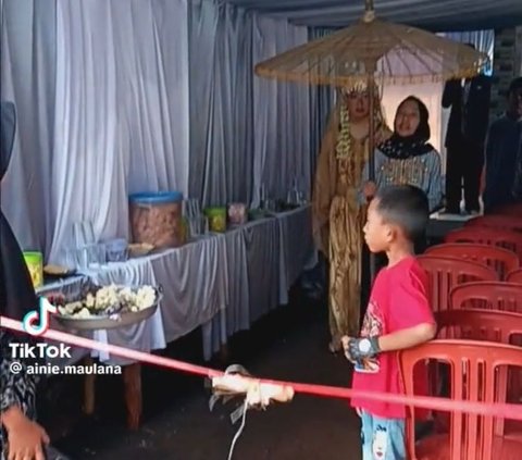 Di saat yang sama, mempelai wanita juga berjalan mendekati pita merah ditemani seorang wanita yang membawa payung.