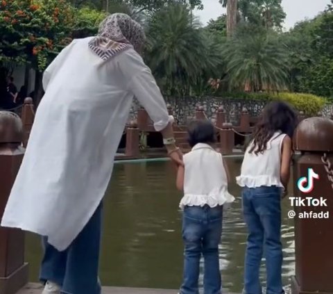 Menemani dua anak balitanya memberi makan ikan di tepi kolam, wanita ini tampak menjaga keduanya. Namun, sang adik yang tampak lebih aktif mencuri perhatian ibunya sehingga sang ibu lebih fokus menjaga anak keduanya.