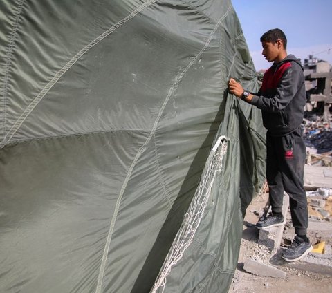 Seorang pria memperbaiki parasut militer yang dimanfaatkan sebagai tenda darurat di atas puing-puing rumahnya di Jalur Gaza, Palestina, pada Rabu (3/4/2024). Parasut tersebut sebelumnya digunakan oleh militer untuk menerjunkan bantuan kemanusiaan melalui udara. AFP