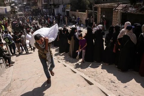 Seorang pria Palestina membawa sekantong tepung, sementara yang lain menunggu di depan titik distribusi yang didirikan oleh Save Youth Future Society (SYFS), yang bekerja sama dengan Program Pangan Dunia PBB (WFP), di Kota Gaza pada 26 Juni 2025. (BASHAR TALEB/AFP)