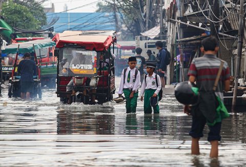 Sejumlah anak dengan seragam sekolah tampak menenteng sepatunya saat melewati banjir rob yang merendam kawasan Muara Baru, Jakarta Utara, Selasa (14/1/2025).