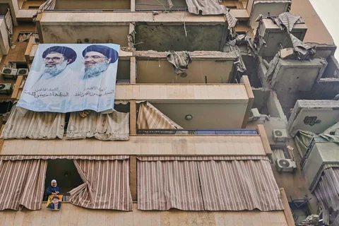 Seorang perempuan memasang spanduk bergambar mendiang Hassan Nasrallah di balkon rumahnya yang rusak, berlatar poster besar Nasrallah dan penggantinya Hashem Safieddine, sehari setelah serangan udara Israel menewaskan kepala staf Hizbullah di Hareit Hreik, Lebanon, Senin (2411/2025).