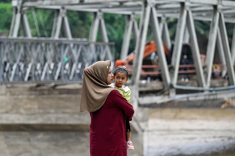 Seorang ibu menggendong anaknya dekat  pembangunan jembatan bailey untuk memperbaiki sementara jembatan Juli yang rusak akibat bencana banjir bandang di Bireuen, Aceh, Minggu (14/12/2025),