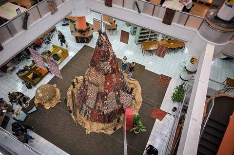 Mahasiswa Indonesia berpose di samping dekorasi Natal sepanjang 63 meter bermotif budaya Papua yang dibuat dengan bantuan AI dan dihiasi Tifa, perisai Asmat, serta ornamen tradisional di Universitas Petra Surabaya, Kamis (18/12/2025).