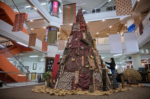 Mahasiswa Indonesia berpose di samping dekorasi Natal sepanjang 63 meter bermotif budaya Papua yang dibuat dengan bantuan AI dan dihiasi Tifa, perisai Asmat, serta ornamen tradisional di Universitas Petra Surabaya, Kamis (18/12/2025).