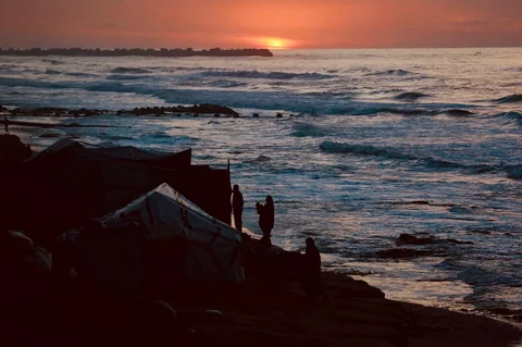 Para pengungsi Palestina berdiri di samping tempat penampungan mereka,saat sunset nam di sebuah pantai di Deir El-Balah di Jalur Gaza tengah,  Selasa (30/12/2025).