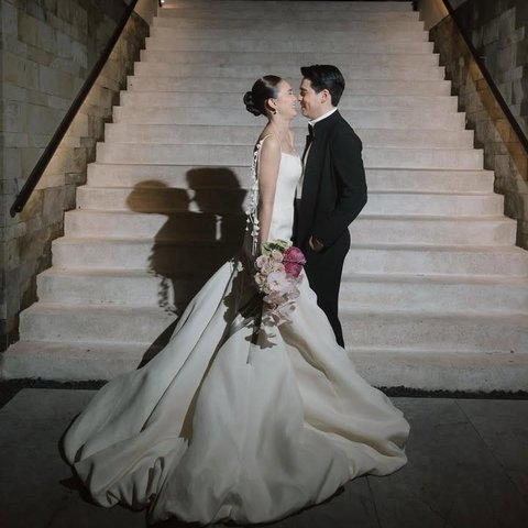 Di resepsi kedua, Luna Maya tampil memukau dalam gaun putih klasik dengan detail pita di punggung dan ekor panjang dramatis. Dipadukan dengan buket bunga pink lembut, penampilannya begitu anggun. Maxime Bouttier tampil gagah dalam tuxedo hitam, berdiri berhadapan mesra di tangga mewah sambil saling melempar senyum penuh cinta. Momen ini menciptakan suasana intimate yang hangat dan romantis.