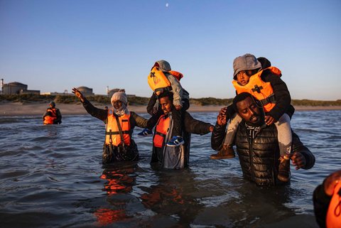 Para migran menggendong anak-anak mereka saat berjalan di air untuk menaiki perahu penyelundup dalam upaya menyeberangi Selat Inggris di lepas pantai Gravelines, Prancis utara, (16/06/2025)