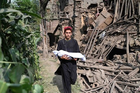 Seorang anak menggendong jenazah seorang kerabatnya terbungkus kain kafan saat berjalan melewati sebuah rumah yang rusak akibat gempa bumi di desa Mazar Dara, Distrik Nurgal, Provinsi Kunar, Afghanistan Timur, Senin (01/09/2025).