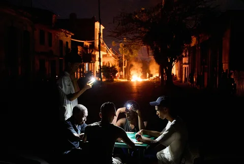 Orang-orang menerangi diri mereka dengan ponsel sambil bermain domino sementara api yang dinyalakan oleh warga yang memprotes pemadaman listrik berkepanjangan berkobar di sebuah jalan di Havana, Kuba, Kamis, Kamis malam (14/05/2026).