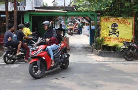 Pengendara melintas di samping spanduk peringatan dan larangan melintas di perlintasan liar jalur kereta api di kawasan Tebet, Jakarta, Jumat(15/05/2026).