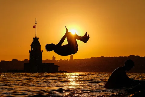 Orang-orang melompat ke Selat Bosporus dengan Menara Maiden sebagai latar belakang saat matahari terbenam di Istanbul, Turki, Minggu (28/06/2026).