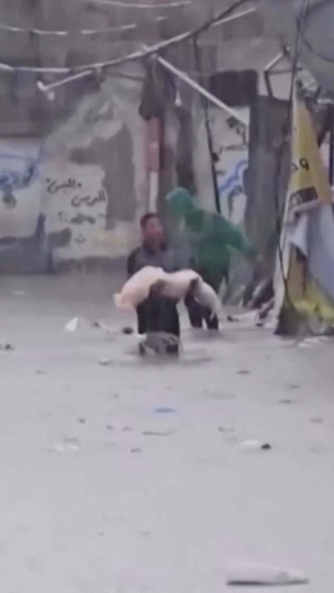 Seorang pria Palestina berjalan di tengah banjir dan hujan deras dengan menggendong jenazah seorang anak korban serangan Israel di Jabalia, Jalur Gaza Utara, pada 13 Desember 2023. Anas Al-Shareef/via REUTERS<br>