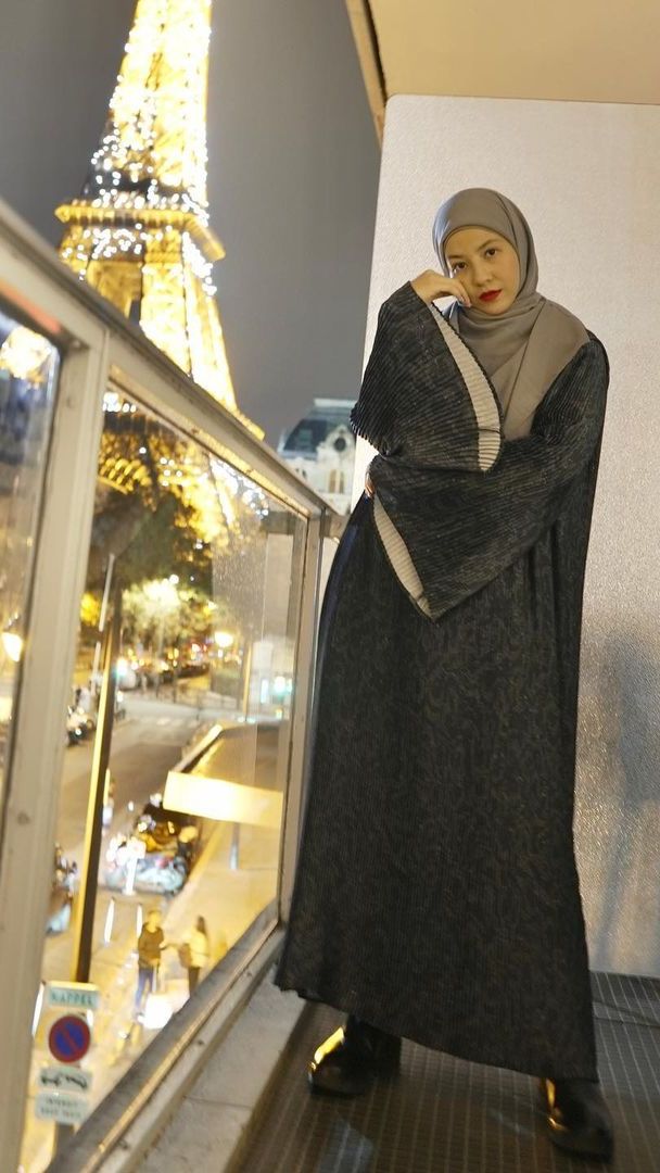 Dia mengenakan dress hitam dengan corak hijau tua dipadukan dengan hijab berwarna abu-abu.