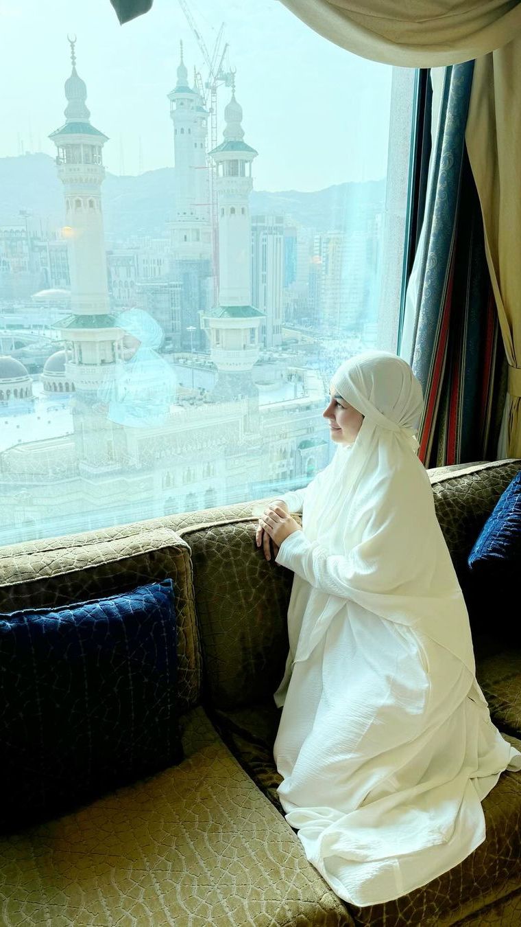 Nisya tampak berpose di jendela kamar hotelnya yang menghadap langsung ke masjidil haram.